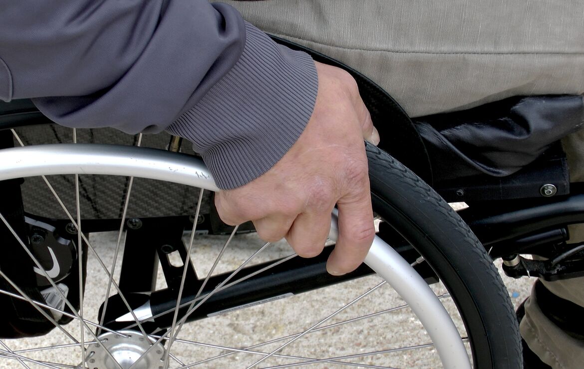 Nahaufnahme von Hand einer Person im Rollstuhl sitzend. Die Hand liegt auf dem Rollstuhlrad auf.