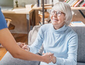 Eine lächelnde Seniorin sitzt auf einem grauen Sofa. Sie hält die Hände einer Angehörigen, die ihr beim Aufstehen hilft.