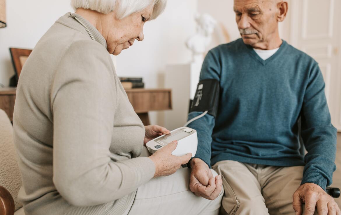 Bild von einem Seniorenpaar, die gegenseitig Blutdruck messen. Der Mann, rechts sitzend, trägt die Manschette. Die Frau, links sitzend, liest das Ergebnis vom zugehörigen Gerät ab.