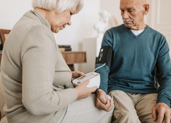 Bild von einem Seniorenpaar, die gegenseitig Blutdruck messen. Der Mann, rechts sitzend, trägt die Manschette. Die Frau, links sitzend, liest das Ergebnis vom zugehörigen Gerät ab.