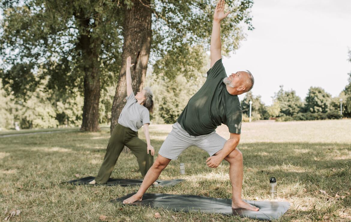 Ein Seniorenpaar ist in einem Park. Beide stehen auf Yogamatten und machen Fitnessübungen.