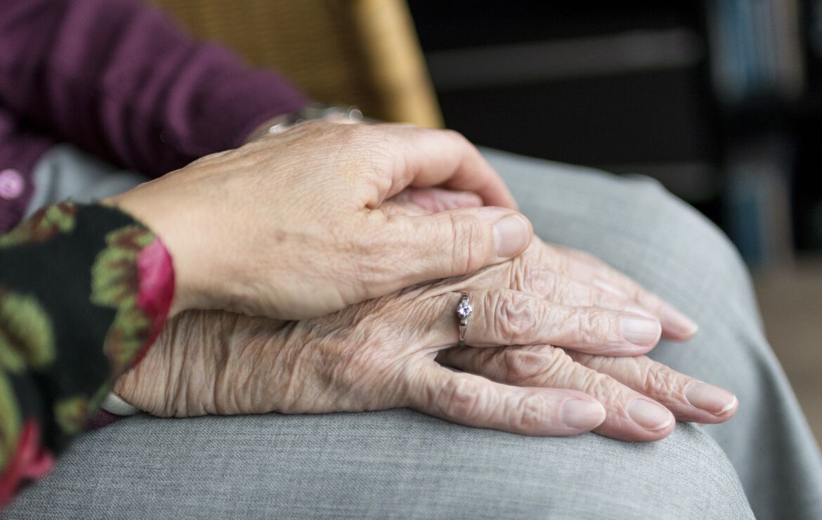 Bildausschnitt von zwei Händen. Eine jüngere Hand liegt Zuneigung spendend auf der Hand einer Seniorin auf.