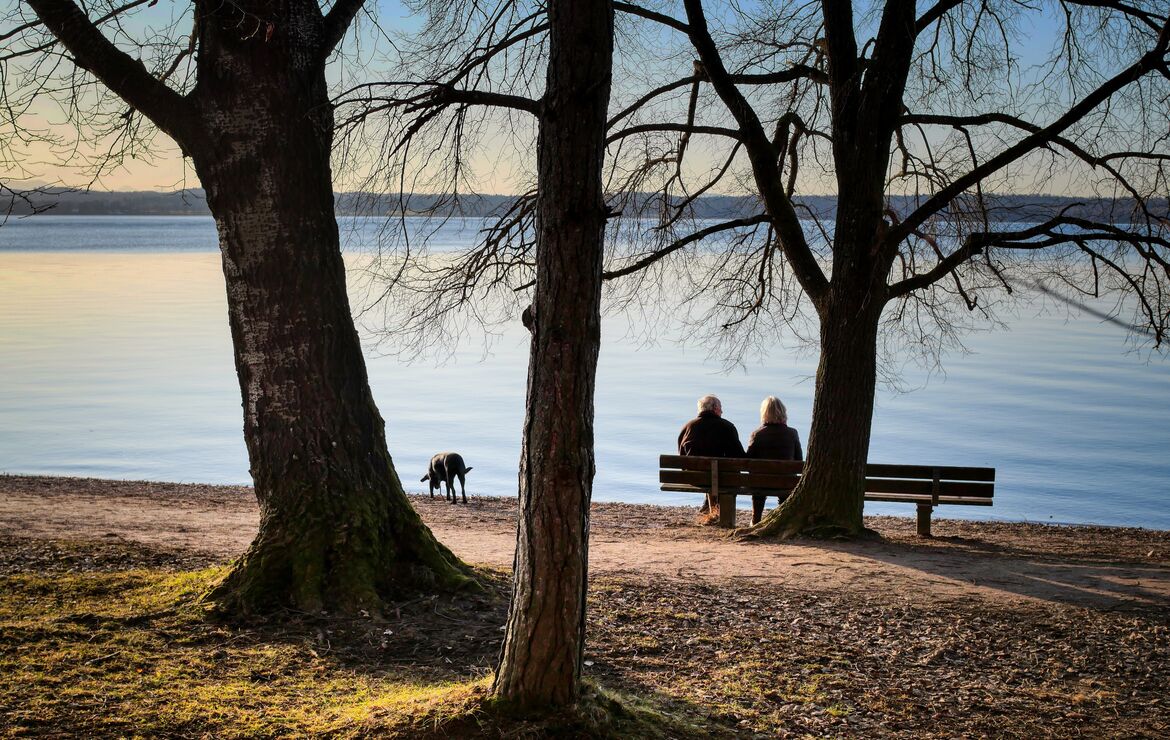 Blick auf ein Paar, das an einem See auf einer Bank sitzt. In kurzer Entfernung erkundet ein Hund das Ufer.