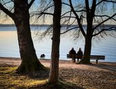 Blick auf ein Paar, das an einem See auf einer Bank sitzt. In kurzer Entfernung erkundet ein Hund das Ufer.