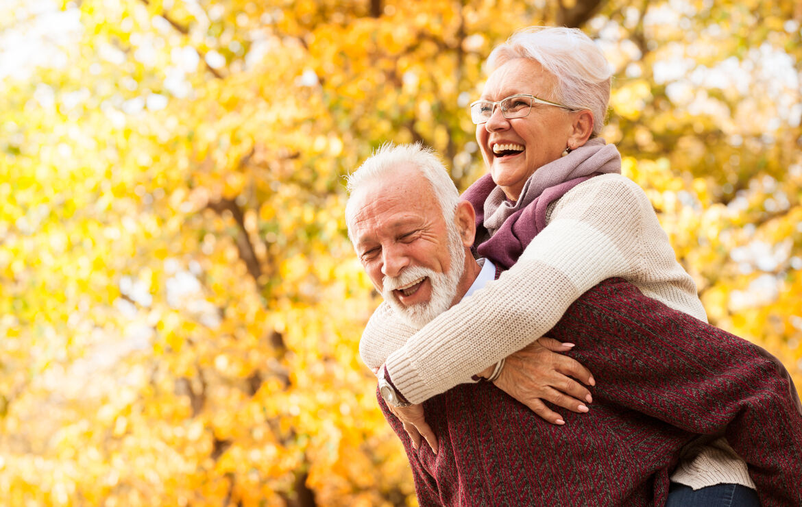 Ein Seniorenpaar genießt gemeinsam den Herbst draußen. Er trägt sie Huckepack. Beide lachen.