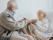 Senior reicht seiner Partnerin, die in einem Pflegebett liegt, ein aufgeschlagenes Buch. Beide lächeln.