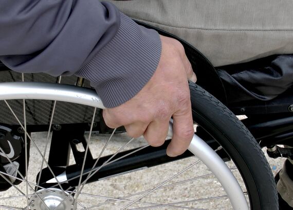 Nahaufnahme von Hand einer Person im Rollstuhl sitzend. Die Hand liegt auf dem Rollstuhlrad auf.