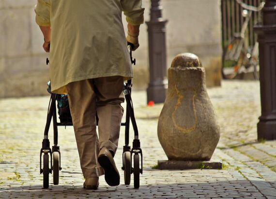 Foto von einer Person, von hinten aufgenommen. Die Person geht an einem Rollator über eine Kopfsteinpflasterstraße.