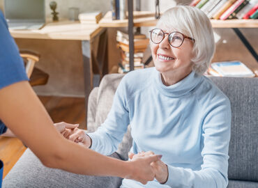 Eine lächelnde Seniorin sitzt auf einem grauen Sofa. Sie hält die Hände einer Angehörigen, die ihr beim Aufstehen hilft.