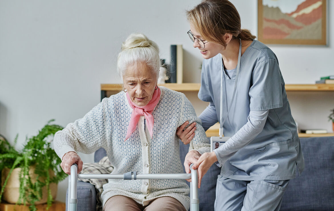 Eine Seniorin sitzt auf einem grauen Sofa. Neben ihr steht leicht gebückt eine Krankenpflegerin , die ihr beim Aufstehen hilft. Die Seniorin ist auf eine Gehhilfe gestützt.