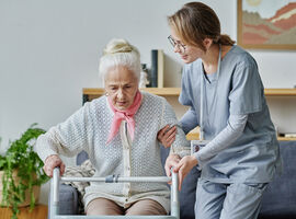 Eine Seniorin sitzt auf einem grauen Sofa. Neben ihr steht leicht gebückt eine Krankenpflegerin , die ihr beim Aufstehen hilft. Die Seniorin ist auf eine Gehhilfe gestützt.