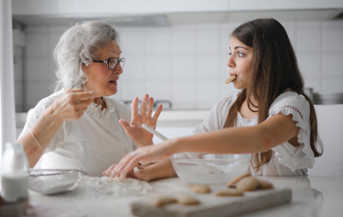 Eine ältere Frau, links im Bild, sitzt mit ihrer Enkelin, rechts im Bild, in der Küche, umgeben von Backutensilien und frisch gebackenen Keksen.