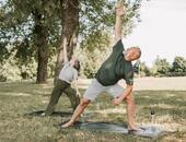 Ein Seniorenpaar ist in einem Park. Beide stehen auf Yogamatten und machen Fitnessübungen.