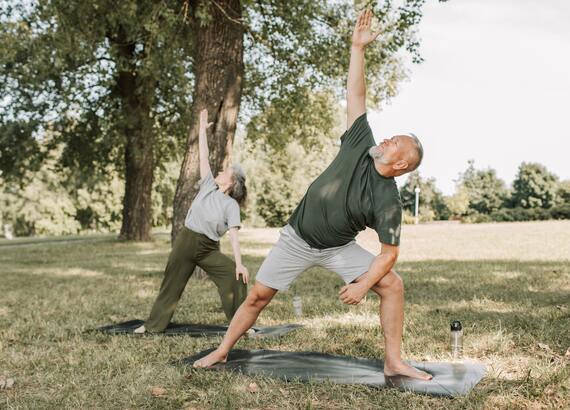 Ein Seniorenpaar ist in einem Park. Beide stehen auf Yogamatten und machen Fitnessübungen.