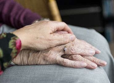 Bildausschnitt von zwei Händen. Eine jüngere Hand liegt Zuneigung spendend auf der Hand einer Seniorin auf.