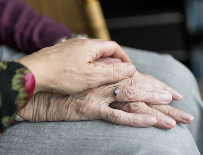 Bildausschnitt von zwei Händen. Eine jüngere Hand liegt Zuneigung spendend auf der Hand einer Seniorin auf.
