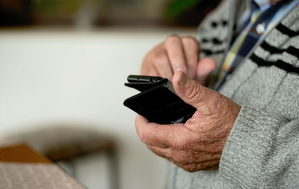 Bildausschnitt von einem älteren Herren, der ein Handy in der Hand hält - der Bildausschnitt zeigt seine Hände mit dem Mobiltelefon.