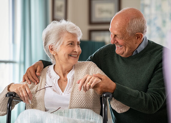 Ein Seniorenpaar in ihrem Wohnzimmer: die Frau sitzt in einem Rollstuhl, der Mann sitzt leicht hinter ihr auf einem Hocker und umfasst sie liebevoll. Sie lächeln einander an.