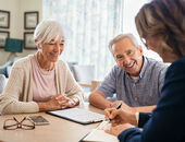 Ein Seniorenpaar sitzt gemeinsam mit einer Pflegeberaterin an einem Tisch. Die Beraterin erklärt etwas anhand von Formularen auf einem Klemmbrett.