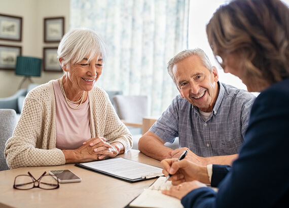 Ein Seniorenpaar sitzt gemeinsam mit einer Pflegeberaterin an einem Tisch. Die Beraterin erklärt etwas anhand von Formularen auf einem Klemmbrett.