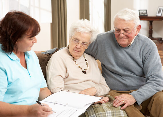 Ein Seniorenpaar sitzt gemeinsam mit einer Altenpflegerin auf der heimischen Couch. Die Altenpflegerin erklärt etwas anhand von Notizen in einem Ordner.