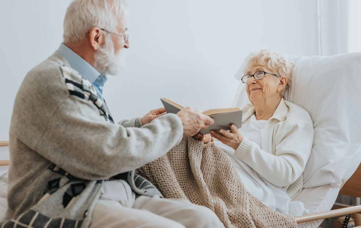 Senior reicht seiner Partnerin, die in einem Pflegebett liegt, ein aufgeschlagenes Buch. Beide lächeln.