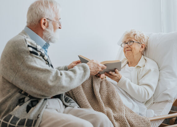 Senior reicht seiner Partnerin, die in einem Pflegebett liegt, ein aufgeschlagenes Buch. Beide lächeln.