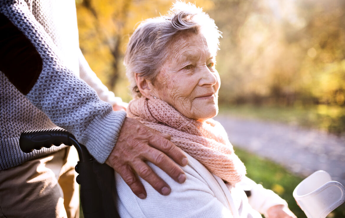 Eine Seniorin, im Rollstuhl sitzend, ist mit einem Angehörigen draußen spazieren. Der Angehörige hat ihr von hinten die Hände auf die Schultern gelegt.