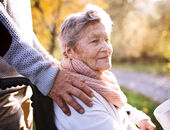 Eine Seniorin, im Rollstuhl sitzend, ist mit einem Angehörigen draußen spazieren. Der Angehörige hat ihr von hinten die Hände auf die Schultern gelegt.