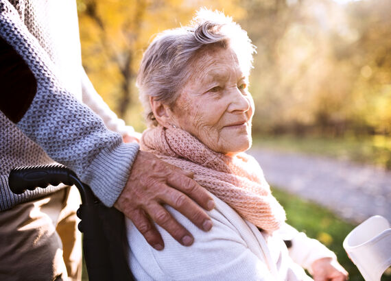 Eine Seniorin, im Rollstuhl sitzend, ist mit einem Angehörigen draußen spazieren. Der Angehörige hat ihr von hinten die Hände auf die Schultern gelegt.