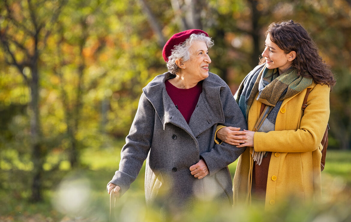 Es wird eine Seniorin beim Spaziergang mit einer Angehörigen gezeigt. Beide sind herbstlich gekleidet. Die Angehörige hat sich bei der Seniorin untergehakt und sie lächeln sich an.