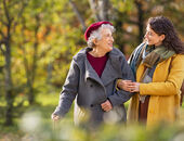 Es wird eine Seniorin beim Spaziergang mit einer Angehörigen gezeigt. Beide sind herbstlich gekleidet. Die Angehörige hat sich bei der Seniorin untergehakt und sie lächeln sich an.