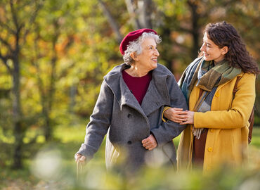 Es wird eine Seniorin beim Spaziergang mit einer Angehörigen gezeigt. Beide sind herbstlich gekleidet. Die Angehörige hat sich bei der Seniorin untergehakt und sie lächeln sich an.
