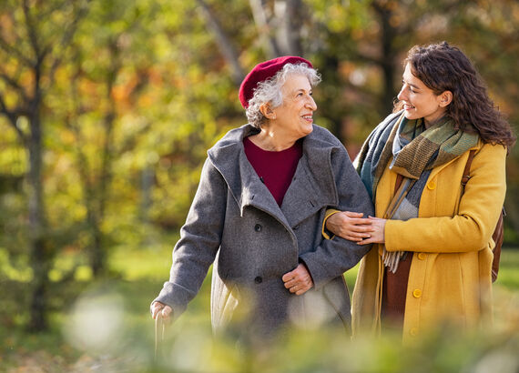 Es wird eine Seniorin beim Spaziergang mit einer Angehörigen gezeigt. Beide sind herbstlich gekleidet. Die Angehörige hat sich bei der Seniorin untergehakt und sie lächeln sich an.