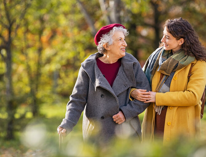 Es wird eine Seniorin beim Spaziergang mit einer Angehörigen gezeigt. Beide sind herbstlich gekleidet. Die Angehörige hat sich bei der Seniorin untergehakt und sie lächeln sich an.