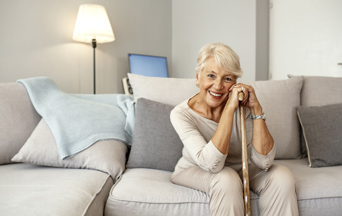 Eine Seniorin sitzt auf einem grau-beigen Sofa. Sie stützt sich mit beiden Händen auf einem Gehstock aus Holz auf und lächelt in die Kamera.