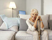 Eine Seniorin sitzt auf einem grau-beigen Sofa. Sie stützt sich mit beiden Händen auf einem Gehstock aus Holz auf und lächelt in die Kamera.