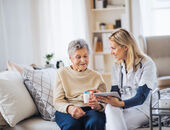 Eine Seniorin und eine Krankenpflegerin sitzen nebeneinander auf einem Bett. Die Pflegerin hält ein Klemmbrett in der einen Hand und reicht der Seniorin mit der anderen Hand eine Medikamentendose. Dabei erklärt sie, wie die Medikamente einzunehmen sind.