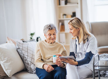 Eine Seniorin und eine Krankenpflegerin sitzen nebeneinander auf einem Bett. Die Pflegerin hält ein Klemmbrett in der einen Hand und reicht der Seniorin mit der anderen Hand eine Medikamentendose. Dabei erklärt sie, wie die Medikamente einzunehmen sind.