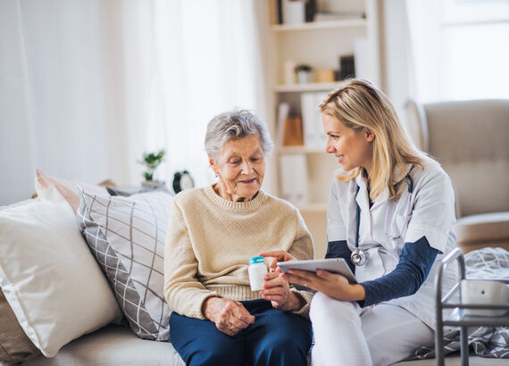 Eine Seniorin und eine Krankenpflegerin sitzen nebeneinander auf einem Bett. Die Pflegerin hält ein Klemmbrett in der einen Hand und reicht der Seniorin mit der anderen Hand eine Medikamentendose. Dabei erklärt sie, wie die Medikamente einzunehmen sind.