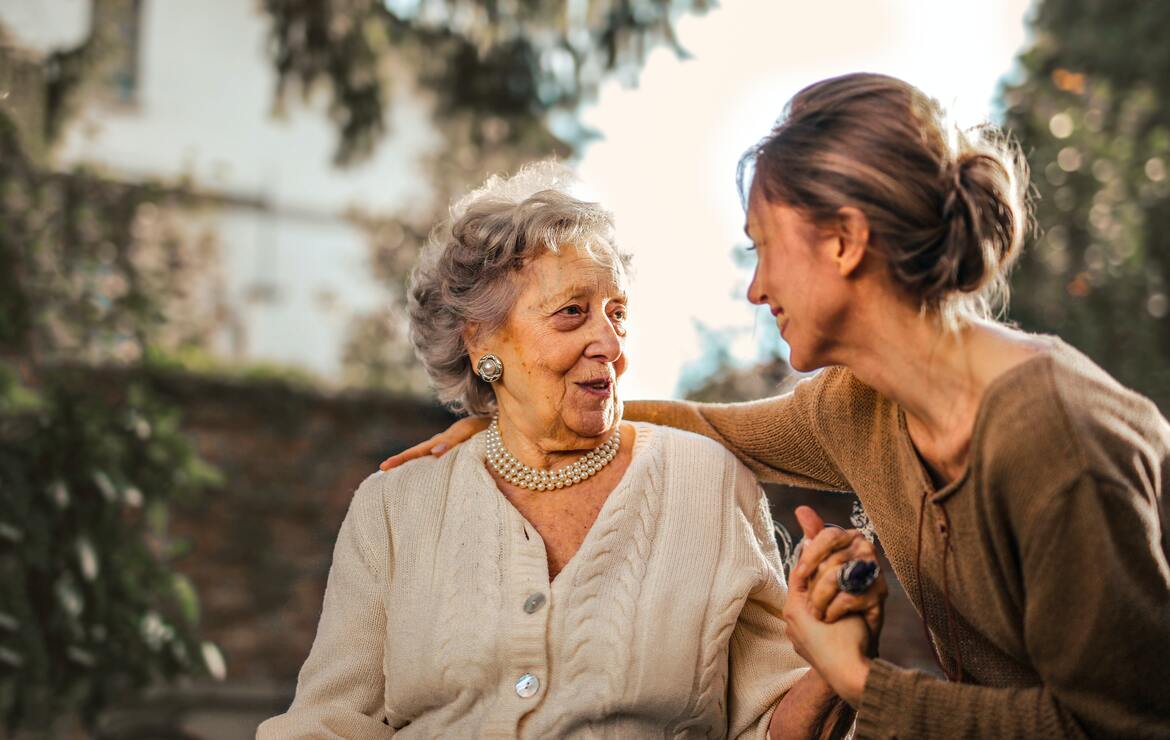 Eine Seniorin, in ihrem Garten sitzend, begrüßt ihre Tochter, die sie besucht.