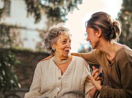 Eine Seniorin, in ihrem Garten sitzend, begrüßt ihre Tochter, die sie besucht.