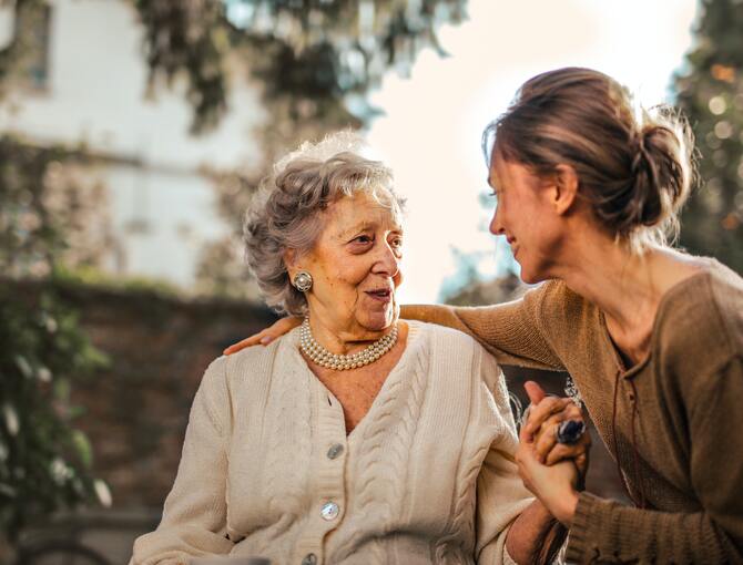 Eine Seniorin, in ihrem Garten sitzend, begrüßt ihre Tochter, die sie besucht.