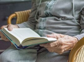 Nachaufnahme einer Seniorin, in einem Rattanstuhl sitzend, die ein Buch liest.